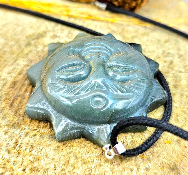 A labradorite carved metal sun pendant with a smiling face and pointed rays, hanging from a black cord. The pendant has a textured, brushed metallic finish in a silver-gray tone in upside down view-My Aura Gemstone