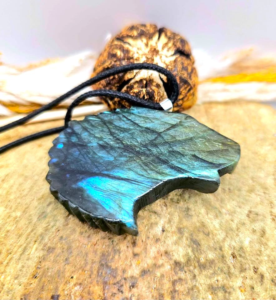 A carved eagle head pendant with a black cord, featuring a metallic gold beak and forehead transitioning to a blue-green patina across the feathers, resting on a textured stone surface in back view-My Aura Gemstones