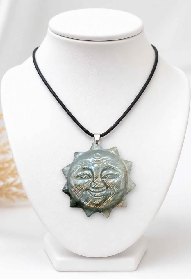 A labradorite carved metal sun pendant with a smiling face and pointed rays, hanging from a black cord. The pendant has a textured, brushed metallic finish in a silver-gray tone displays on jewelry stand-My Aura Gemstones