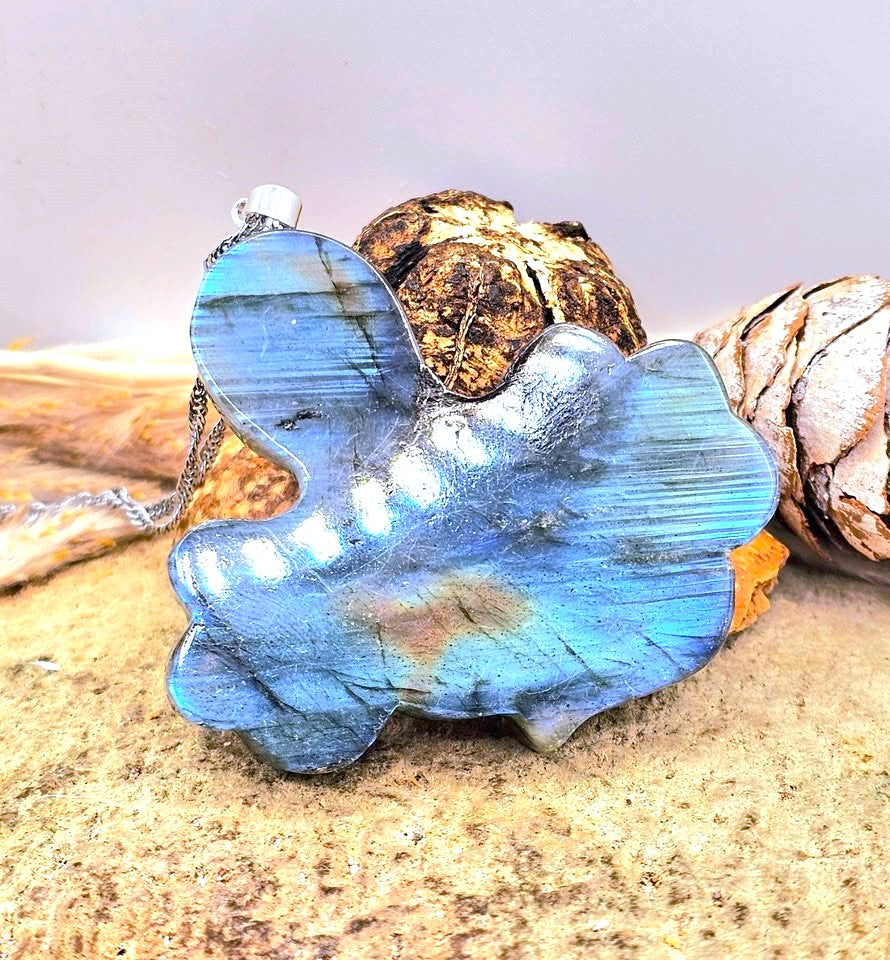 A blue labradorite stone pendant with a silver bail, resting on a sandy surface with dried pinecones in the background.