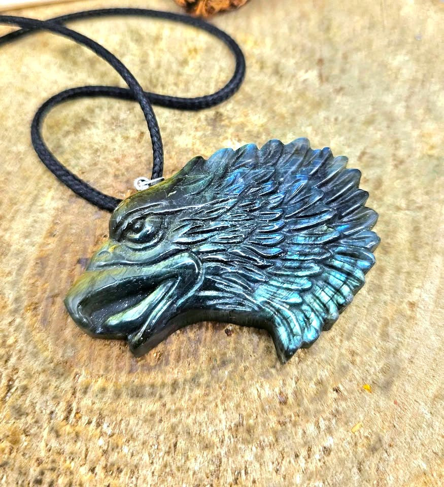 A carved eagle head pendant with a black cord, featuring a metallic gold beak and forehead transitioning to a blue-green patina across the feathers, resting on a textured stone surface-My Aura Gemstones