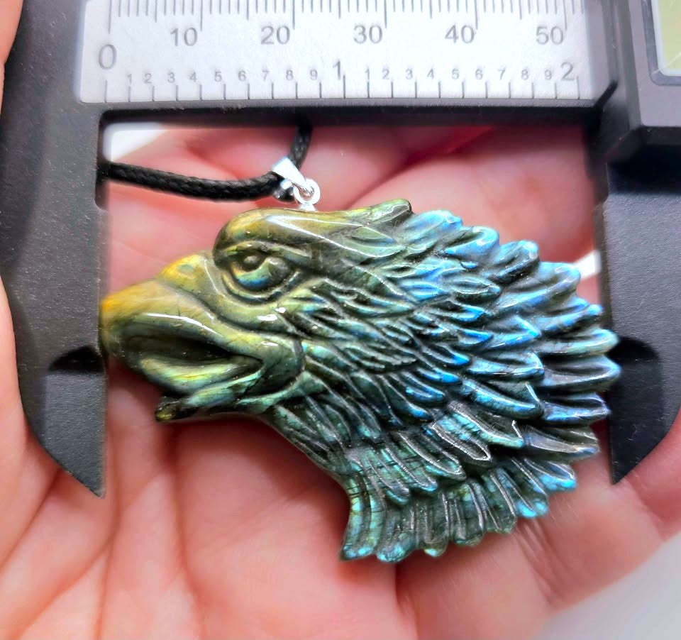 A close-up of an eagle head pendant held in a hand under a caliper, showing a polished iridescent blue-green surface with yellow highlights and a black cord.