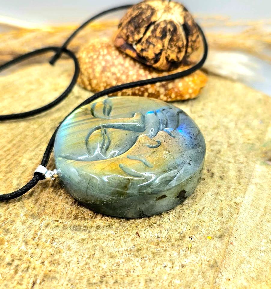 A carved round pendant necklace featuring a serene face design, made from a blue-green stone with iridescent shifts, hanging from a black cord on a sandy surface with decorative shells in the background.