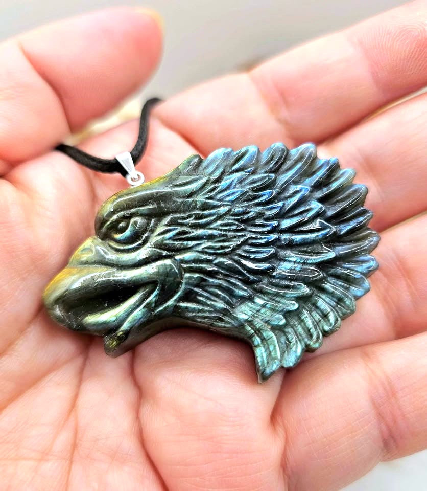 A carved eagle head pendant with a black cord, featuring a metallic gold beak and forehead transitioning to a blue-green patina across the feathers, resting  on hand-My Aura Gemstones