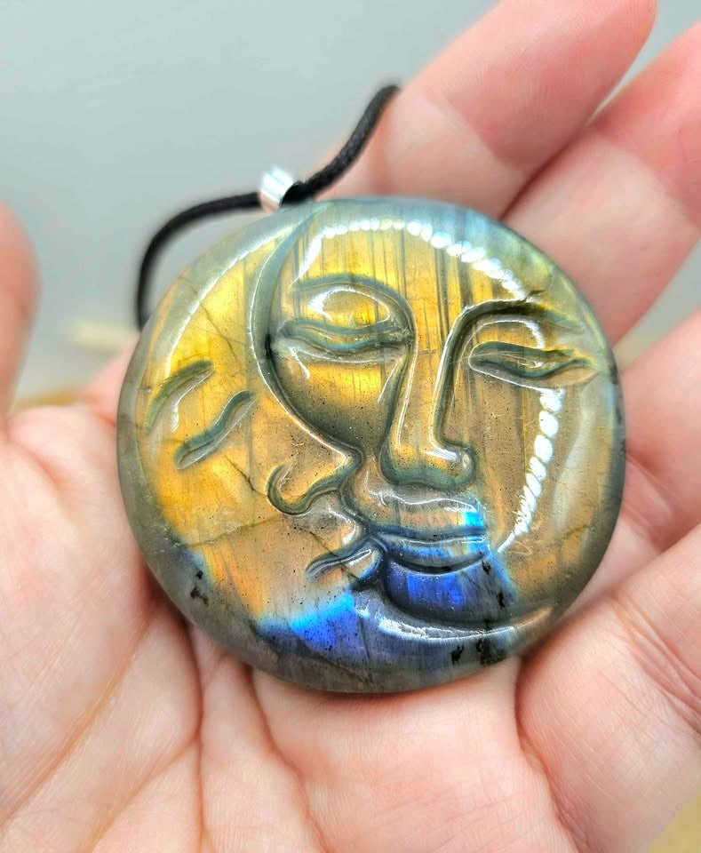 A hand holds a round labradorite pendant carved with a serene face, showing a shimmering gold, green, and blue iridescent finish on a black cord.