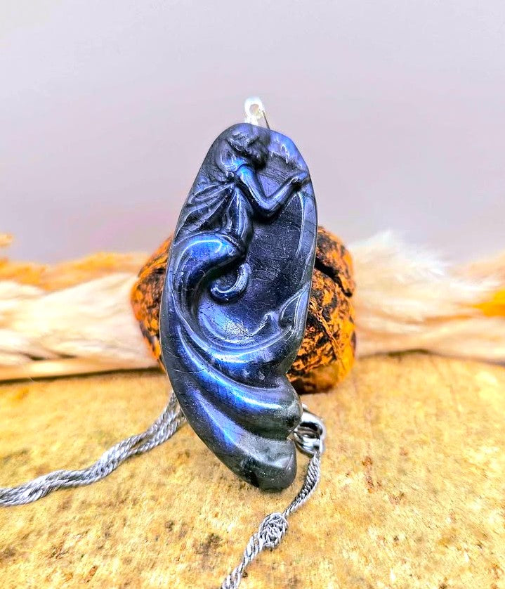 A carved labradorite pendant featuring a mermaid design, hanging from a silver chain against a rustic background of wood and dried grass in front view-My Aura Gemstones