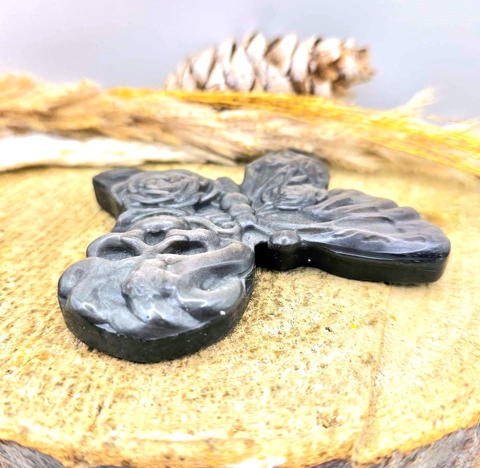 A dark gray to black carved stone cross pendant with floral and skull motifs, displayed on a textured beige wood slice with dried grass and a pinecone in the background.