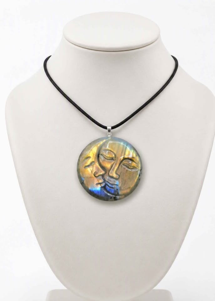 A round pendant necklace displayed on a white mannequin bust. The pendant features a stylized face design with gold, blue, and green iridescent tones, suspended from a black cord.