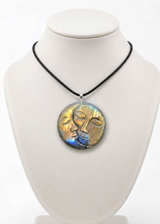 A round pendant necklace displayed on a white mannequin bust. The pendant features a stylized face design with gold, blue, and green iridescent tones, suspended from a black cord.