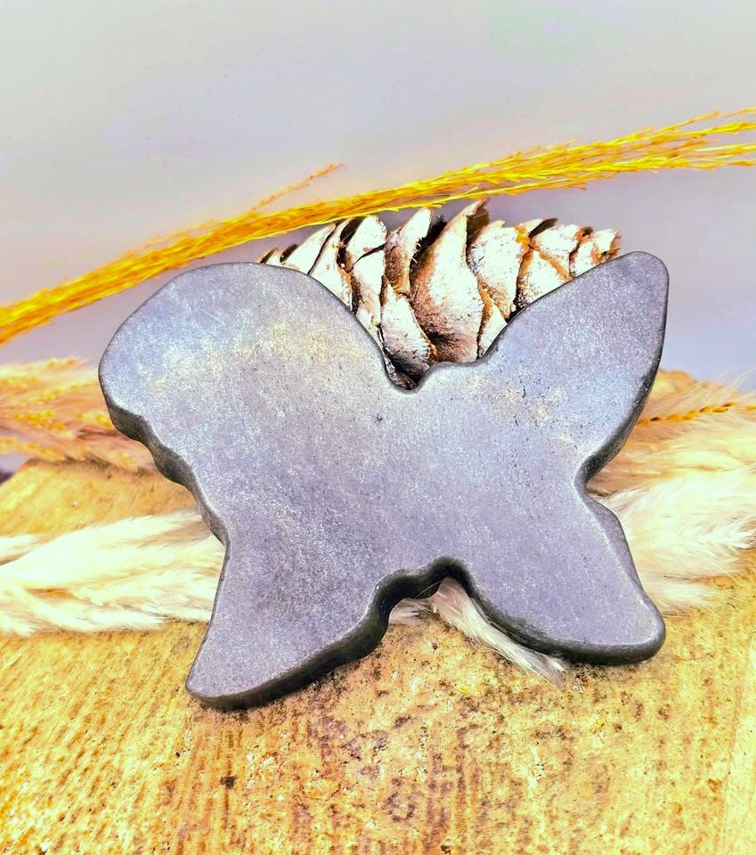 A butterfly-shaped ring made from a dark gray stone, displayed on a piece of wood with a pinecone and dried grass.