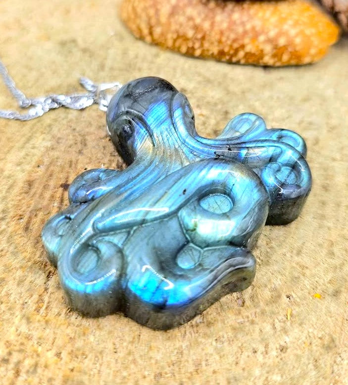A polished labradorite octopus pendant with vibrant blue and green iridescent flashes, hanging from a silver chain on a sandy surface.