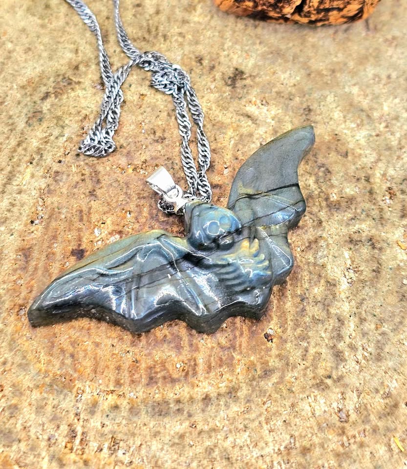 A close-up of a handmade pendant necklace featuring a carved bat-shaped piece made from layered green and blue stone with black veins, attached to a silver chain, resting on a textured tan stone surface.