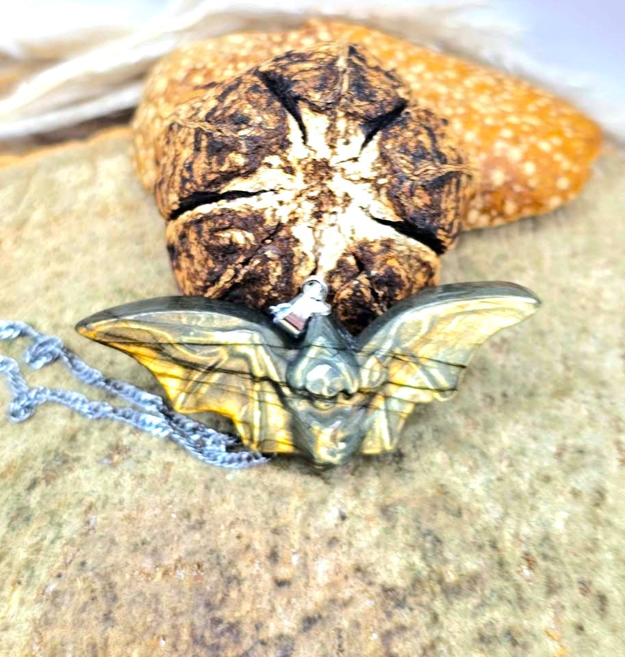 A close-up of a handmade pendant necklace featuring a carved bat-shaped piece made from layered green and blue stone with black veins, attached to a silver chain, resting on a textured tan stone surface.