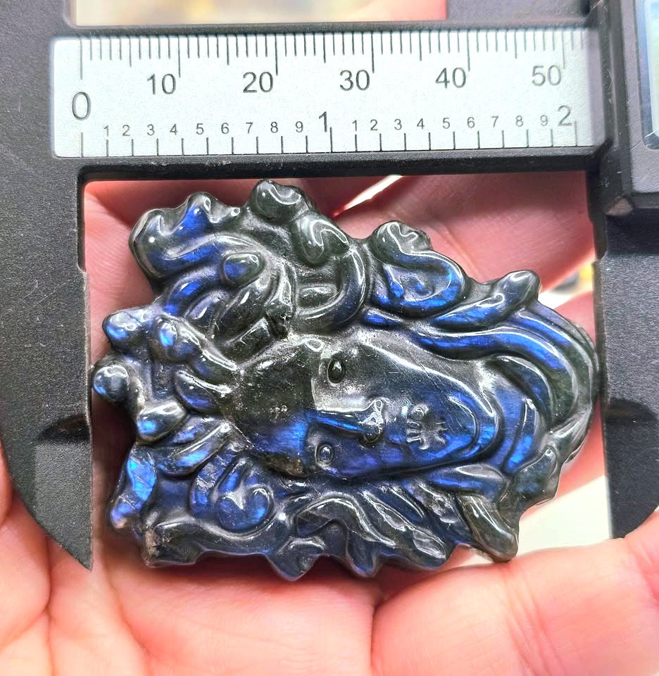A close-up of a carved blue labradorite pendant held in a hand, with a caliper at the top showing its dimensions. The pendant features a stylized fish design with swirling wave-like patterns and small Chinese characters on the side.