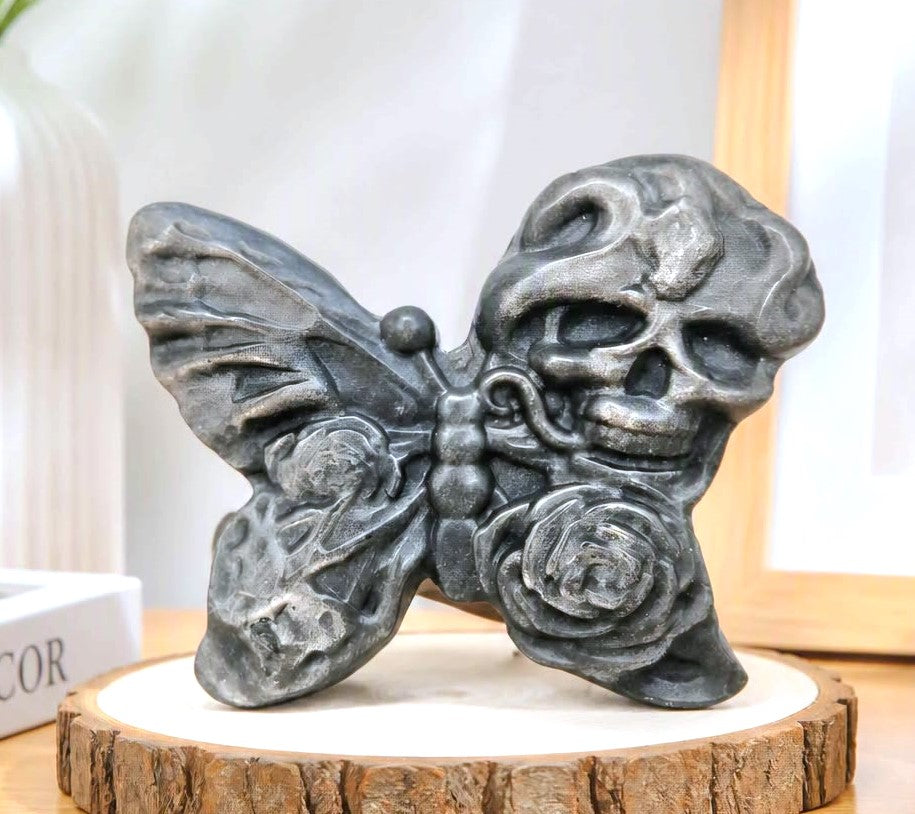 A dark gray gothic silver obsidian stone slice carved with butterfly with a skull on one wing and a rose on the other with indoor desk in background