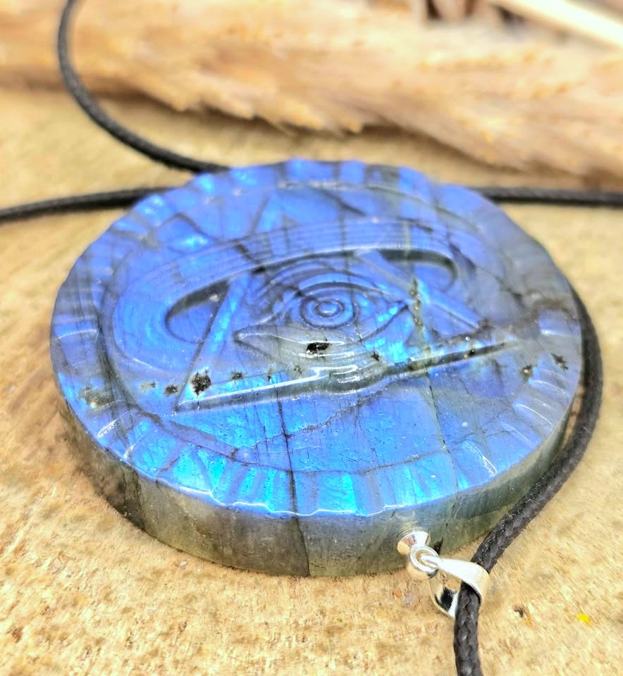 A round labradorite pendant necklace featuring an eye of providence design, showcasing a vibrant blue and green iridescent sheen, suspended from a black cord with a silver clasp on a textured tan background.