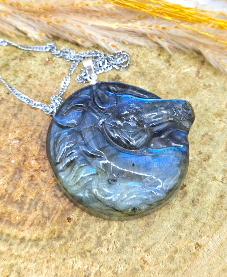 A round labradorite pendant necklace featuring a carved horse head design, resting on a textured stone surface with dried wheat stalks in the background.