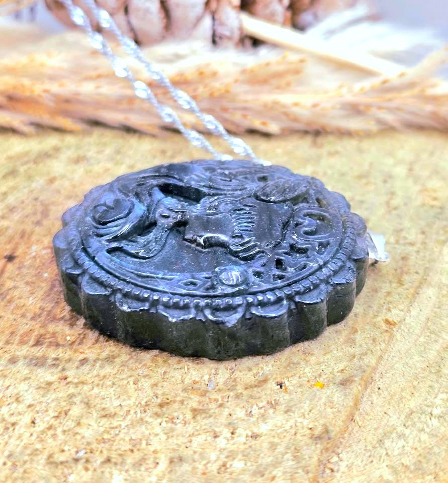 A round black carved pendant necklace with a silver-toned chain, resting on a textured tan surface with dried wheat stalks blurred in the background.