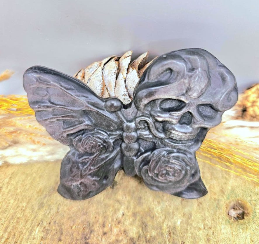 A dark gray gothic silver obsidian stone slice carved with butterfly with a skull on one wing and a rose on the other, resting on a textured surface with dried wheat in top view-My Aura Gemstones