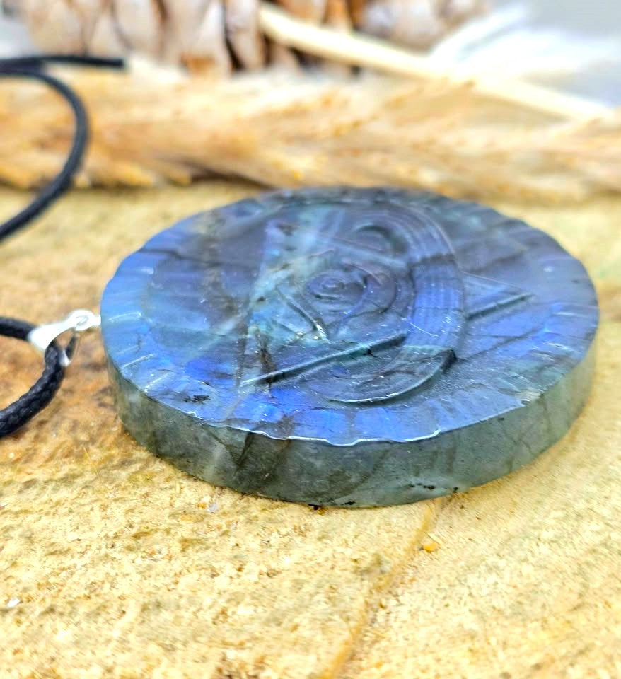 A round labradorite pendant necklace with a carved Viking-style design, resting on a textured stone surface with a black cord and silver clasp visible.