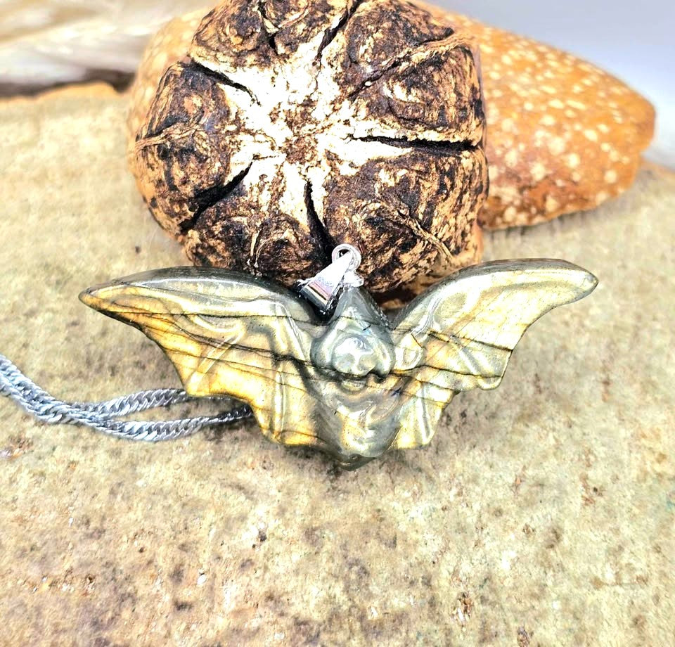 A bat-shaped pendant necklace with a metallic gold and dark gray finish, resting on a textured stone surface with a small silver chain.