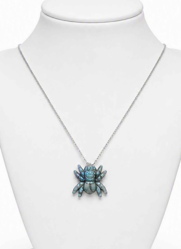 A silver chain necklace with a spider-shaped pendant featuring iridescent blue-green shell-like segments, displayed on a white neck mannequin.