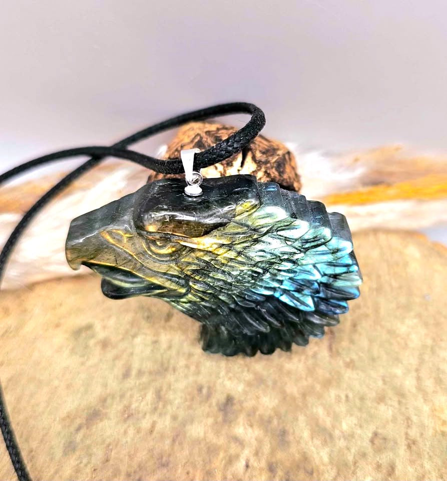 A carved eagle head pendant with a black cord, featuring a metallic gold beak and forehead transitioning to a blue-green patina across the feathers, resting on a textured stone surface in top view-My Aura Gemstones