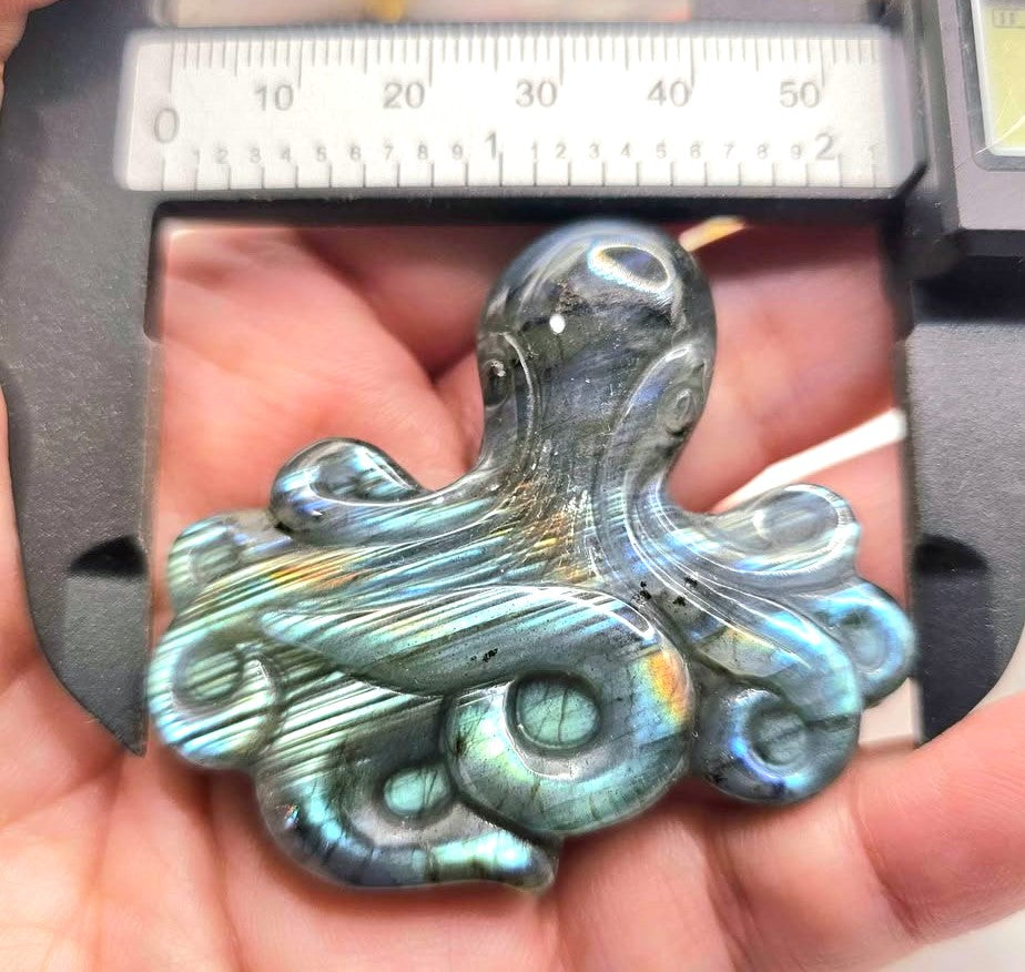 A close-up of a carved octopus-shaped pendant or charm made from iridescent labradorite, held in a person's hand under a metal caliper for scale.