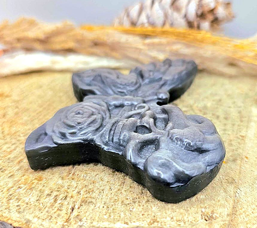 A dark gray carved stone pendant featuring a skull and rose motif, displayed on a textured tan surface with blurred natural background elements.
