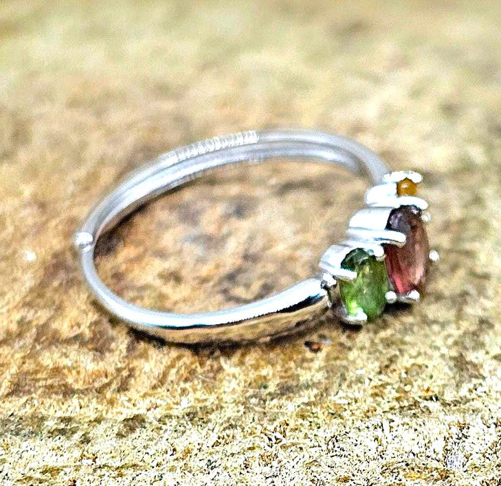 A sterling silver ring with three gemstones (green tourmaline, pink tourmaline, and a small yellow topaz) resting on a textured sandy surface.
