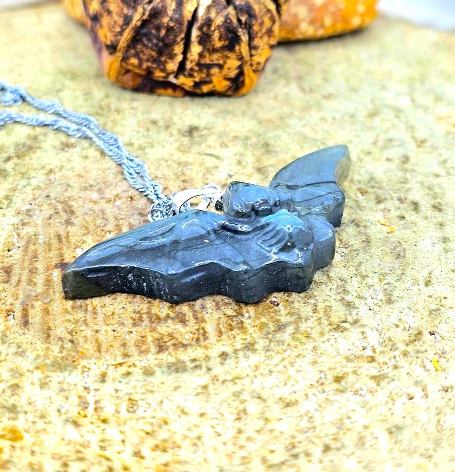 A close-up of a handmade pendant necklace featuring a carved bat-shaped piece made from layered green and blue stone with black veins, attached to a silver chain, resting on a textured tan stone surface.