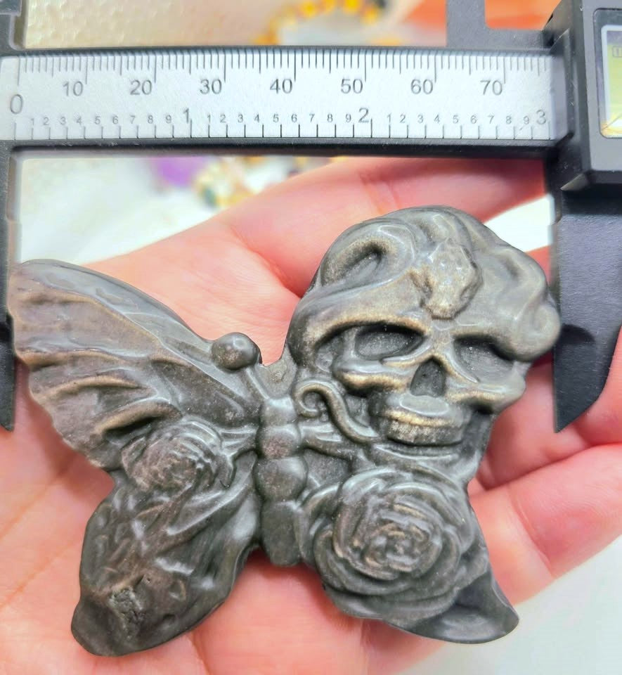 A close-up of a dark metallic butterfly-shaped pendant or charm held in a hand, featuring a skull and rose design. A digital caliper is positioned above it for scale.