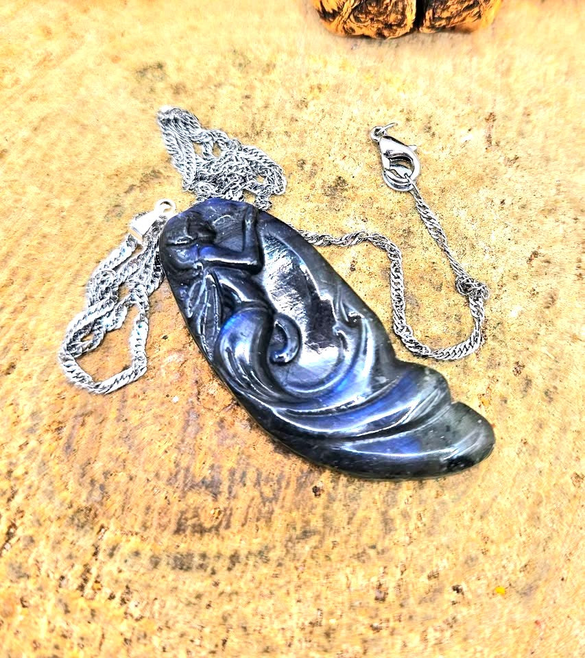 A carved labradorite pendant featuring a mermaid design, hanging from a silver chain against a rustic background of wood and dried grass.-My Aura Gemstones