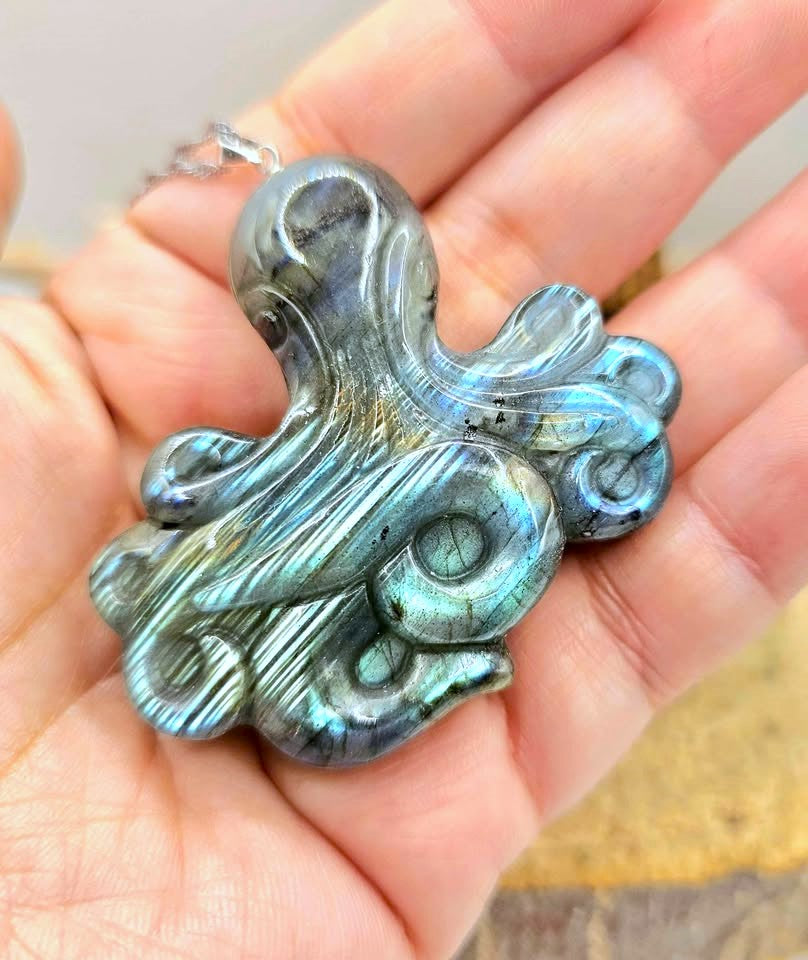 A carved labradorite octopus pendant with a silver bail, held in a person's hand to show scale. The pendant features natural labradorite iridescence with blue, green, and gray tones.