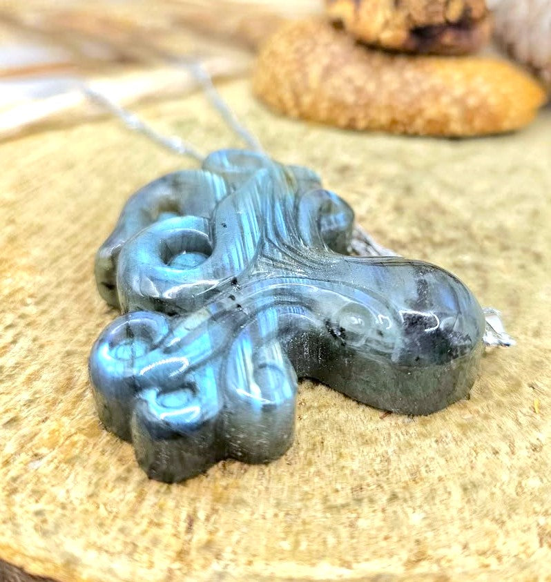 A polished iridescent blue-gray pendant with swirling carved patterns, hanging on a silver chain over a textured tan surface.