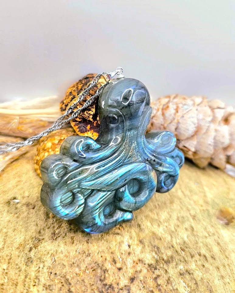 A carved octopus pendant made of iridescent blue-green labradorite stone, hanging from a silver chain and resting on a textured sandy surface with dried grass and a pinecone nearby.