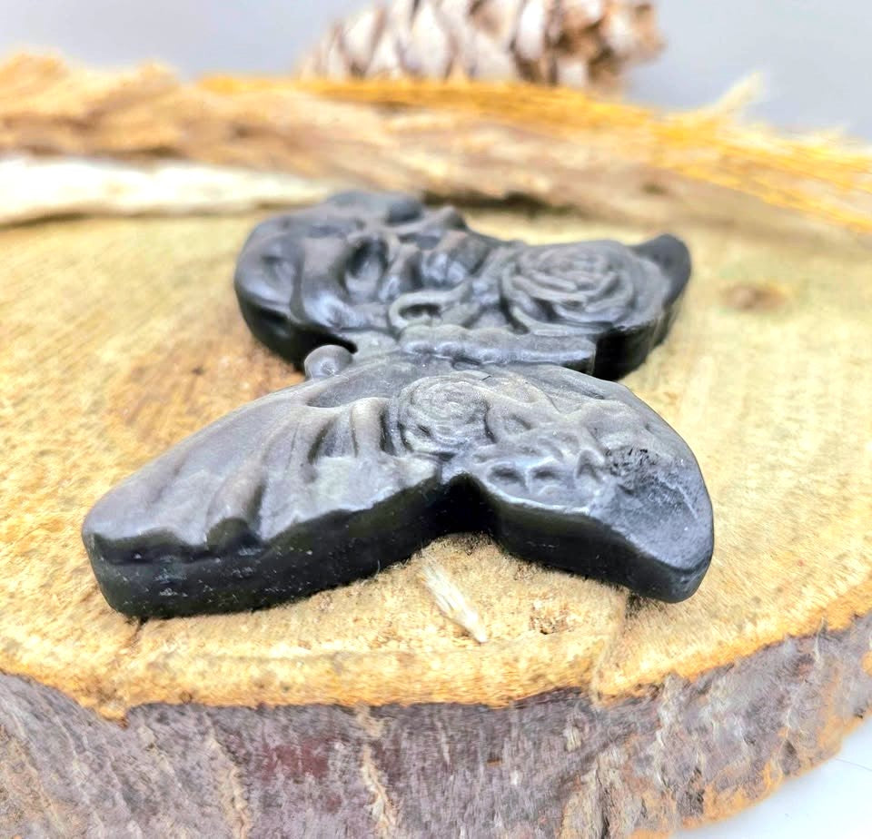 A dark gray gothic silver obsidian stone slice carved with butterfly with a skull on one wing and a rose on the other, resting on a textured surface with dried wheat in side view -My Aura Gemstones