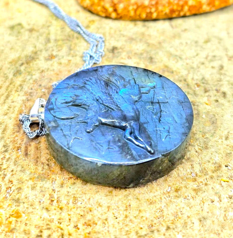 A round labradorite pendant necklace featuring a carved deer design, resting on a sandy surface with a silver chain.