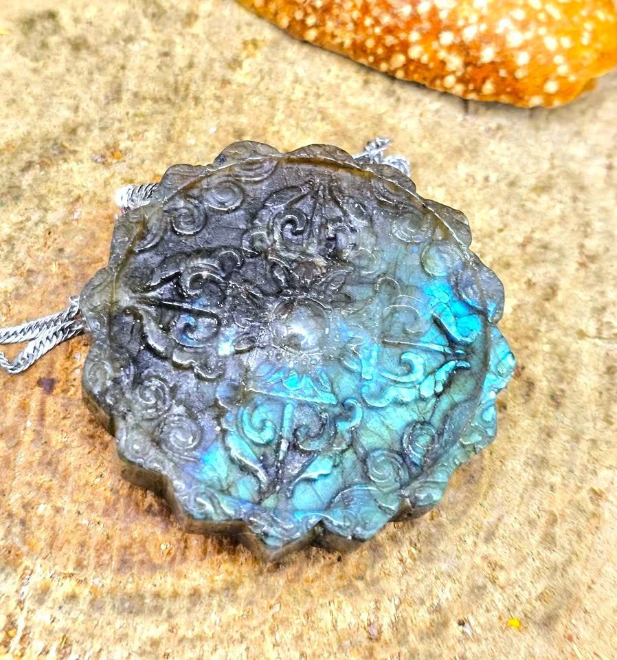 A round labradorite pendant with a silver-toned chain, featuring a swirling floral and scroll pattern. The stone displays a strong blue iridescent flash, set against a textured sandy background.