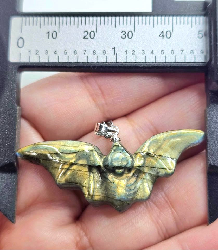 A close-up of a handmade pendant necklace featuring a carved bat-shaped piece made from layered green and blue stone with black veins, attached to a silver chain, resting on a textured tan stone surface.