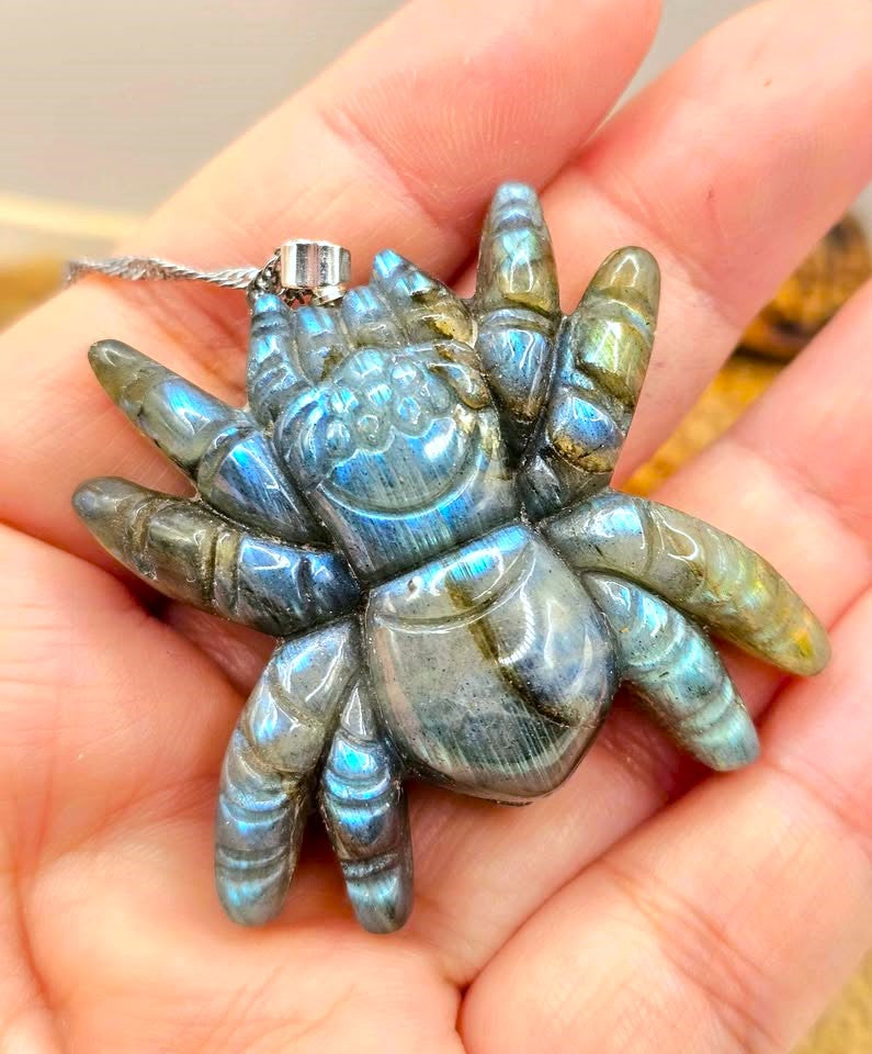A hand holding a carved labradorite spider pendant. The spider features iridescent blue, green, and gray labradorite stone with a silver bail and chain.