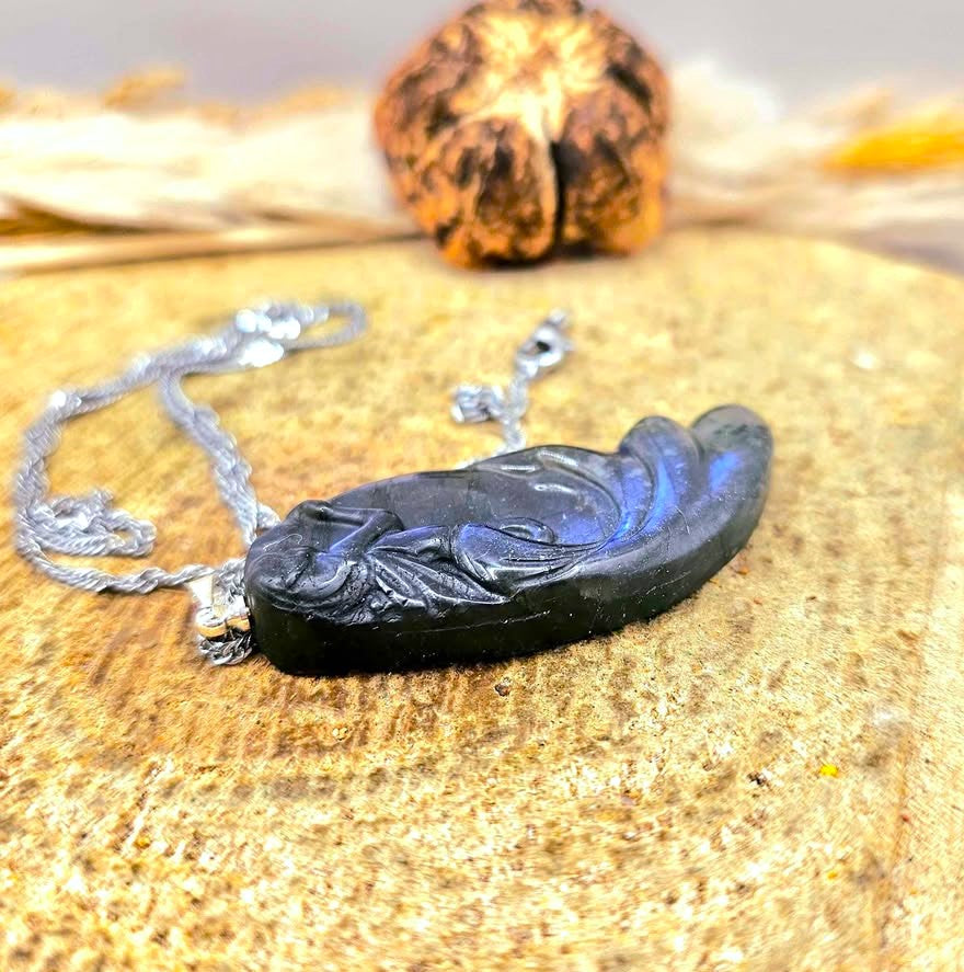 A carved labradorite pendant featuring a mermaid design, hanging from a silver chain against a rustic background of wood and dried grass.-My Aura Gemstones