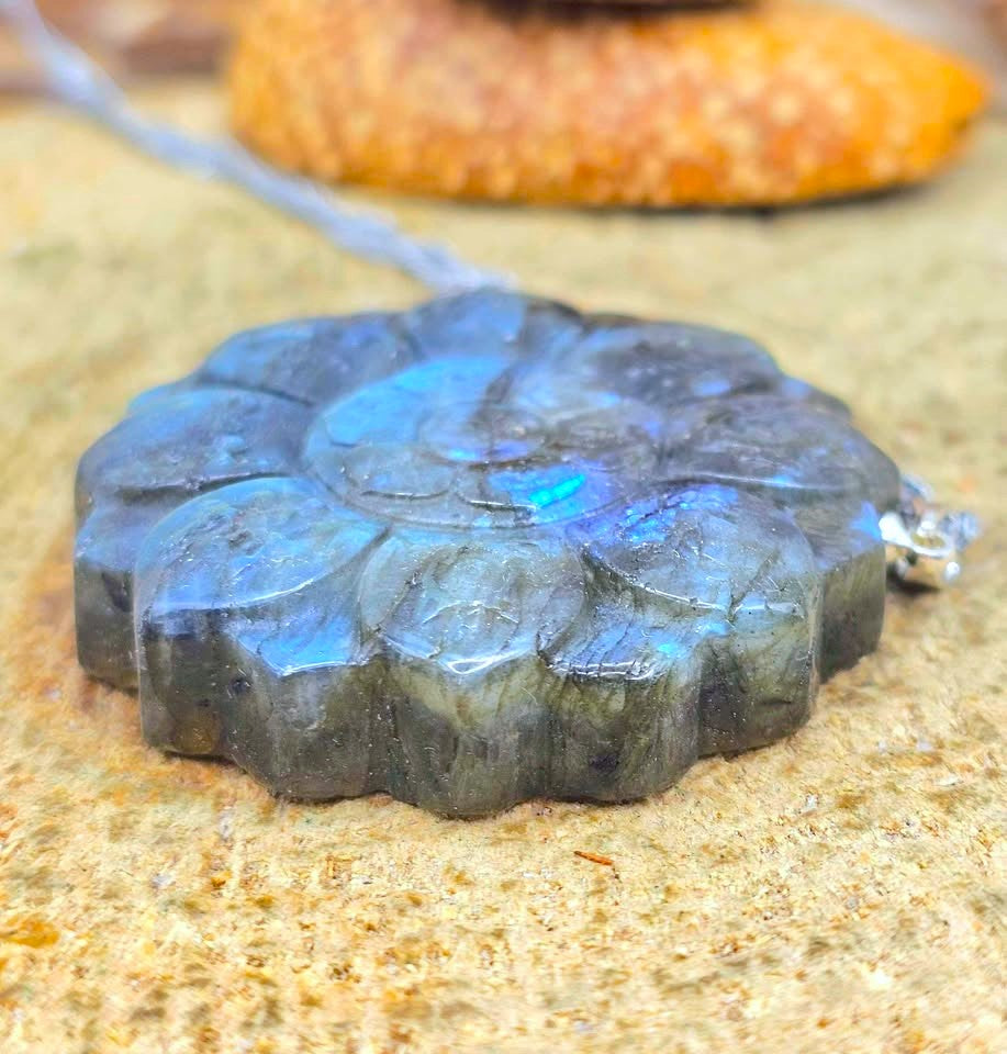 A carved round labradorite pendant with a floral or sunburst pattern, resting on a sandy surface. The pendant displays a characteristic blue iridescent sheen on a dark gray base, with a silver bail and cord visible.