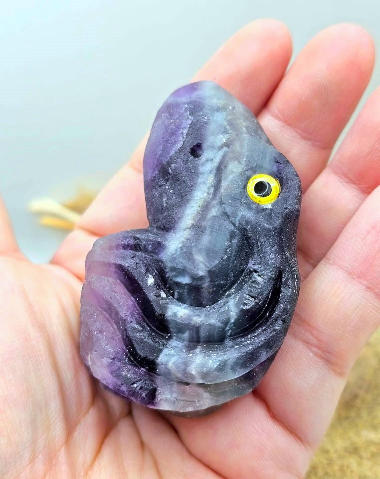 A hand holding a small carved stone or crystal figurine shaped like a stylized face or creature, with a purple and gray marbled appearance and a bright yellow eye detail.