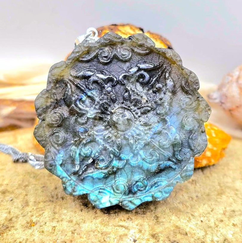 A round, scalloped pendant or medallion with intricate carved floral and scroll patterns, featuring a blue-green iridescent finish over a dark gray base, resting on a sandy surface with small decorative stones.