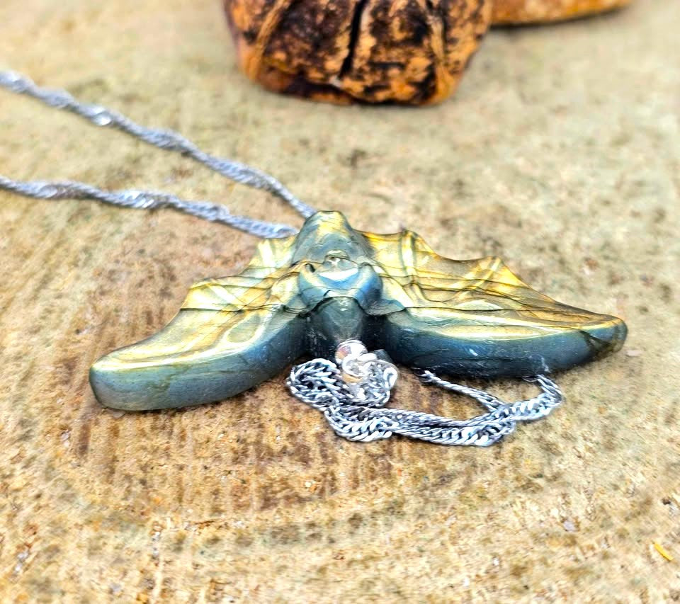 A close-up of a handmade pendant necklace featuring a carved bat-shaped piece made from layered green and blue stone with black veins, attached to a silver chain, resting on a textured tan stone surface.
