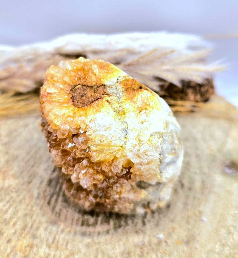 A natural mineral specimen with orange, yellow, and white crystalline texture, displayed on a textured beige surface with a blurred background.