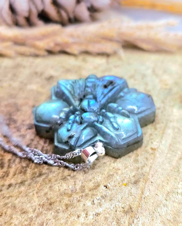 A carved stone pendant necklace with a blue-green iridescent finish and a silver-toned chain, resting on a textured sandy surface.