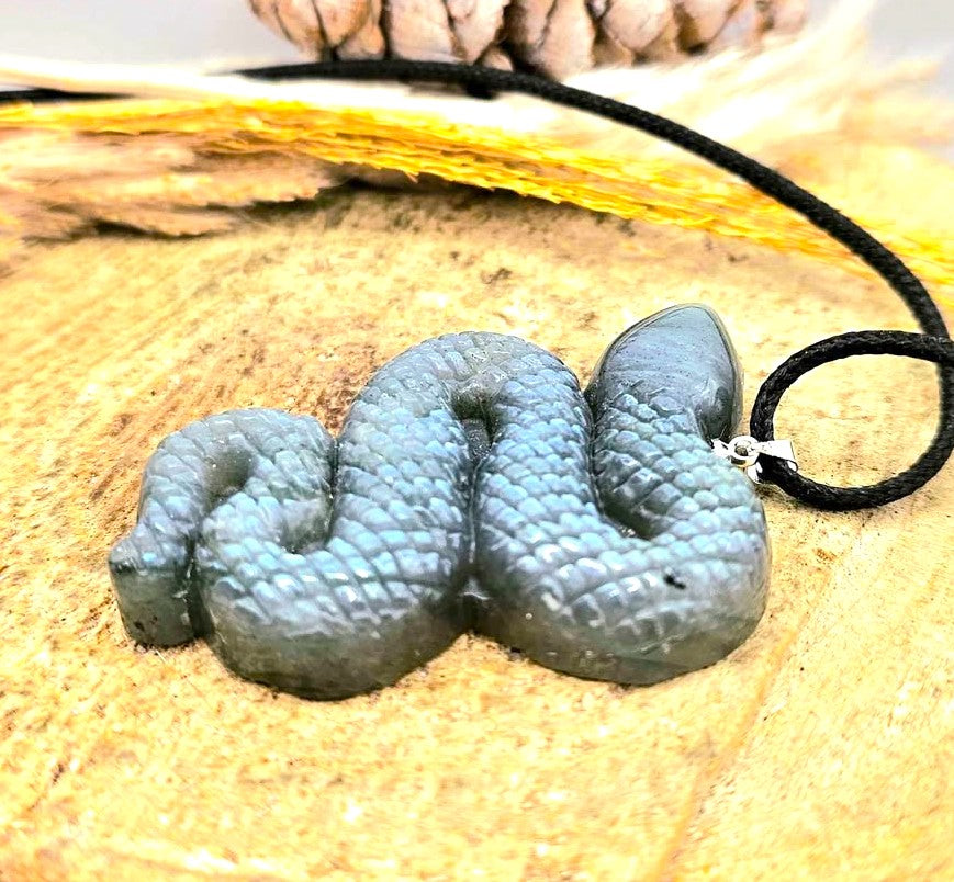 A sculpted snake pendant with a textured, scale-like surface in a muted blue-gray color, attached to a black cord, displayed on a sandy surface.