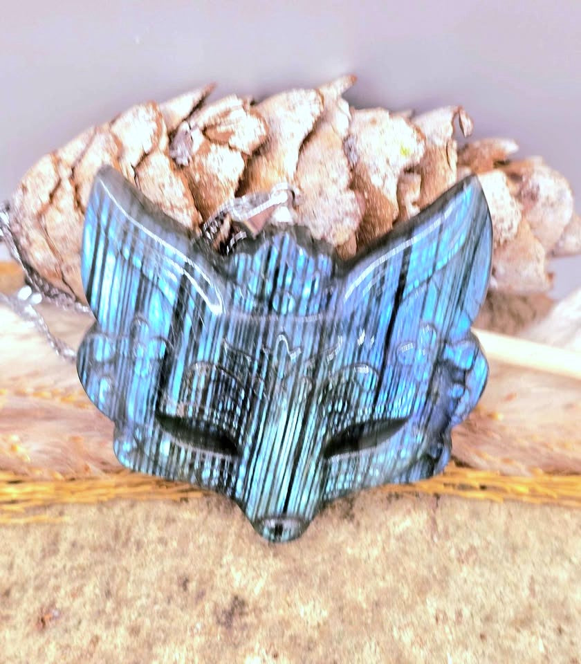 A blue-green iridescent fox-head pendant or charm with engraved details, displayed against a natural background with dried leaves and a tan textured surface.